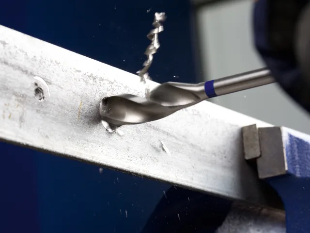 Drill bit drilling into a metal plate with visible chips and a blue glove in the background.