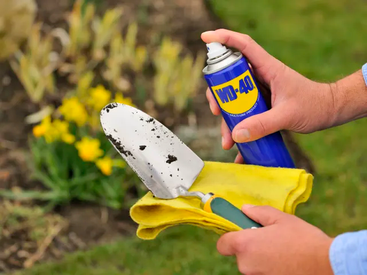Person cleaning a garden trowel with a yellow cloth and WD-40 spray, outdoors.