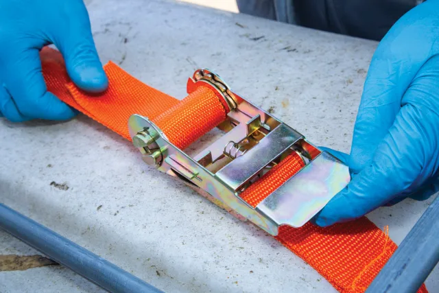 Orange ratchet strap with a metal buckle being adjusted by hands wearing blue gloves on a concrete surface.