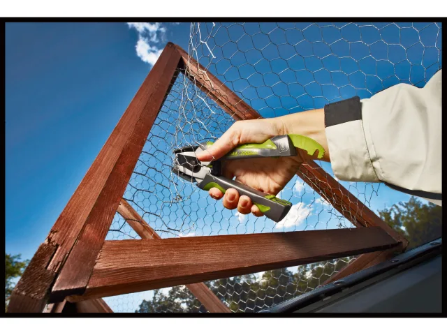 Person using a tool to work on a wooden frame with chicken wire against a blue sky.