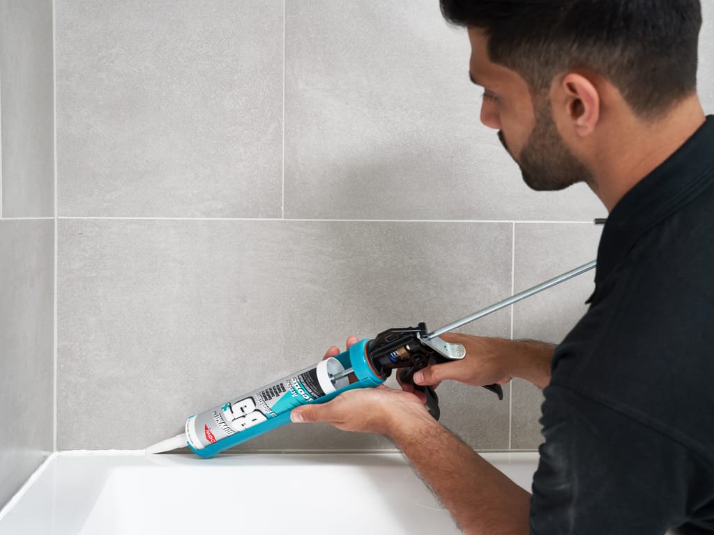 Person applying caulk to a bathroom wall with a caulking gun.