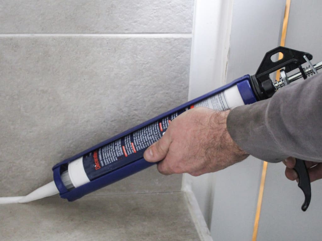 Person using a caulk gun to apply sealant along a bathroom wall.