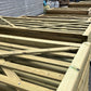 Natural wood garden fence frames and slats stacked outdoors in a workshop or yard area, with sheds and other fencing materials visible in the background.