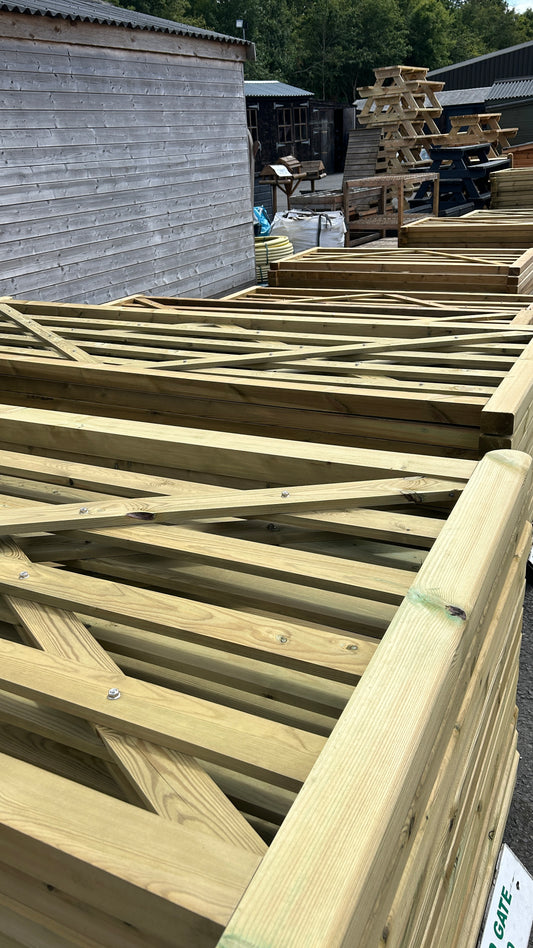 Natural wood garden fence frames and slats stacked outdoors in a workshop or yard area, with sheds and other fencing materials visible in the background.