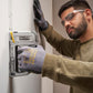 A man wearing safety glasses and work gloves uses a Stanley Power-Tool 18V Cordless Multi-Task Stapler on a white wall.