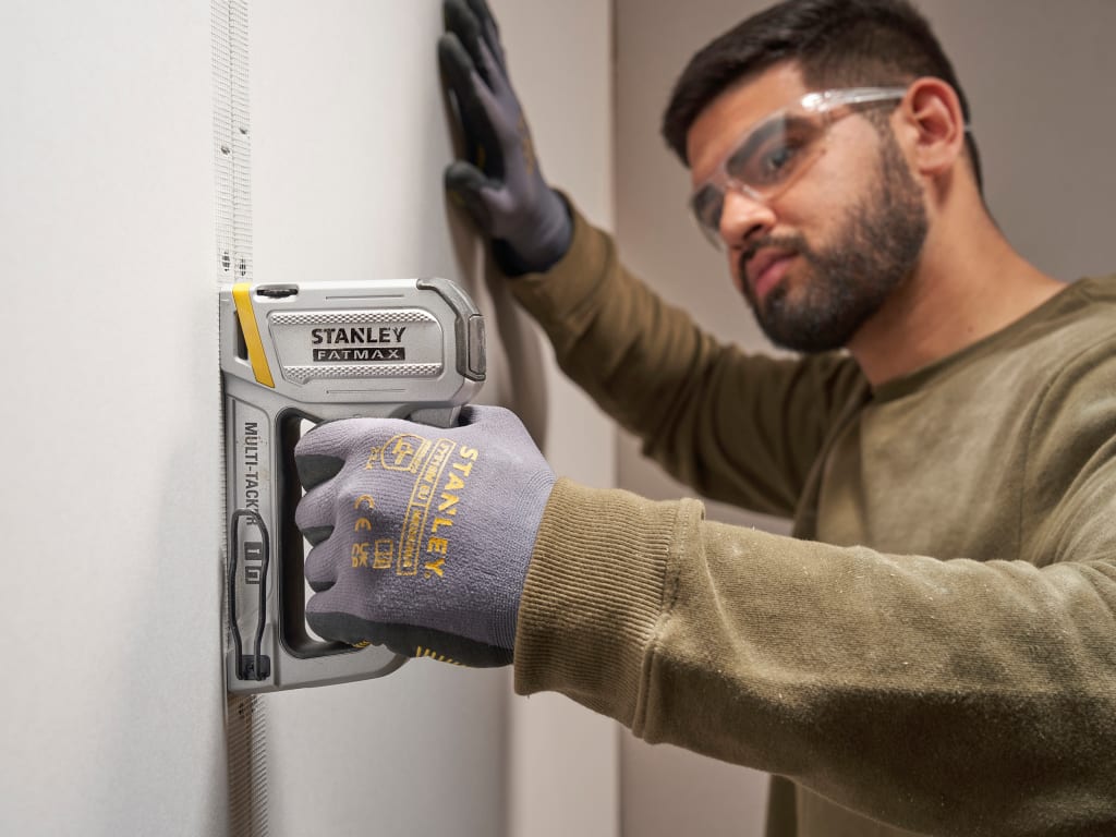 A man wearing safety glasses and work gloves uses a Stanley Power-Tool 18V Cordless Multi-Task Stapler on a white wall.