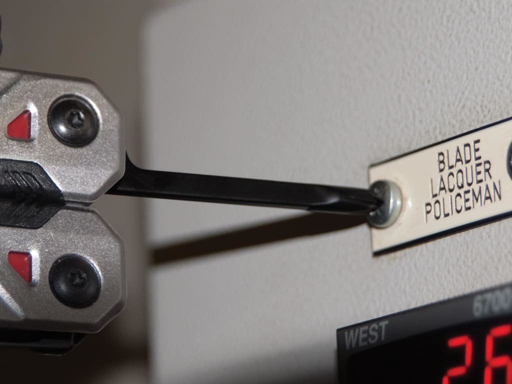 Close-up of a metal tool holding a black rod against a white machine panel with a sign reading 'BLADE LACQUER POLICEMAN' and a red digital display.