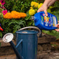 Person pouring a liquid from a blue bottle labeled 'Booja' into a watering can with flowers in the background.