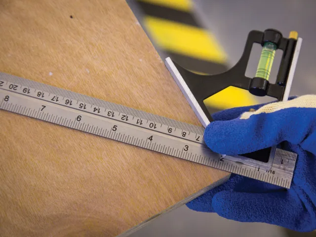 Hand wearing a blue glove using a metal ruler on a wooden surface with a blurred background.