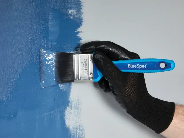 Hand wearing a black glove holding a BlueSpot brand paintbrush applying blue paint to a surface.