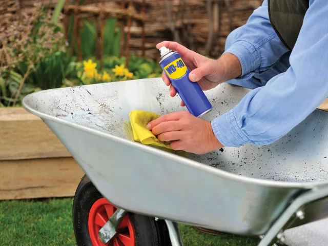 Person cleaning a wheelbarrow with a spray bottle and yellow cloth outdoors.