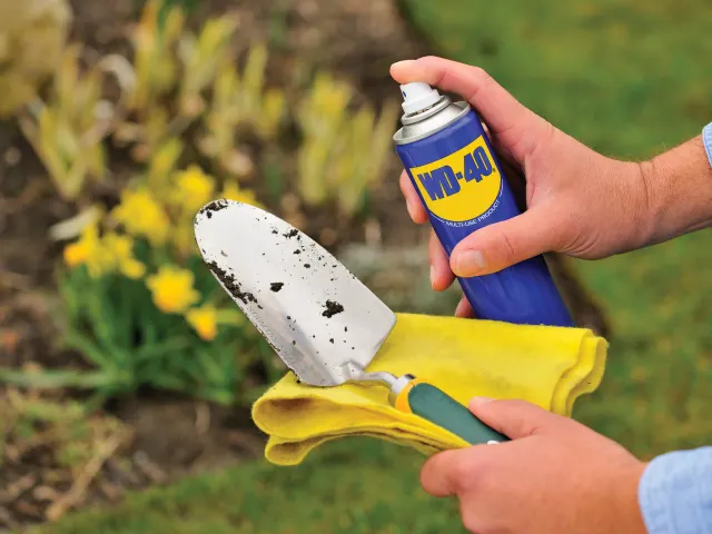 Person cleaning a garden trowel with a yellow rag and WD-40 spray in a garden setting.