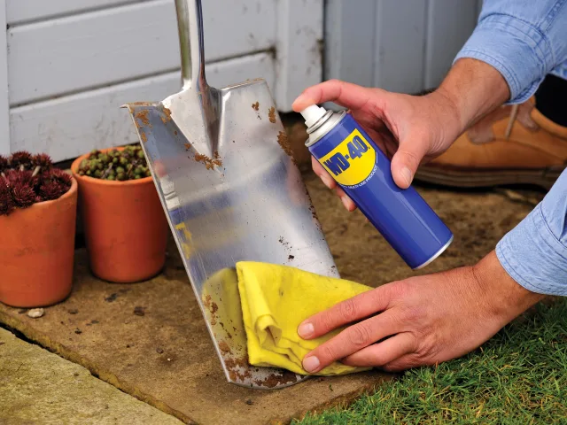 Person cleaning a rusty metal shovel with a yellow cloth and WD-40 spray.