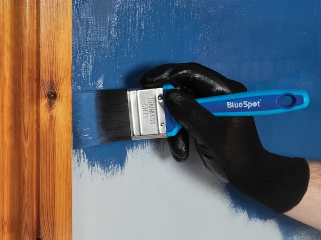 Hand wearing a black glove holding a BlueSpot paintbrush against a blue surface.
