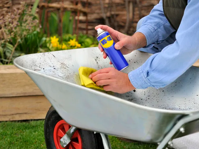Person cleaning a wheelbarrow with a spray bottle and yellow cloth outdoors.