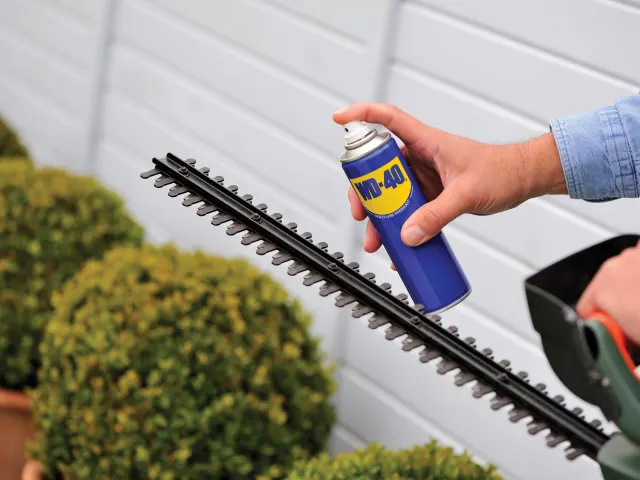 Person using a spray can labeled 'W240' on a hedge trimmer against a white wall.