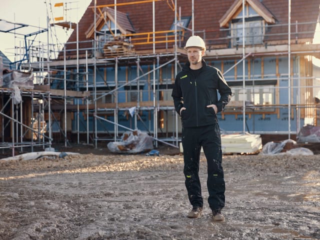 Person in a hard hat and black outfit standing on a construction site with scaffolding and a building in the background.