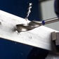 Drill bit drilling into a metal plate with visible chips and a blue glove in the background.