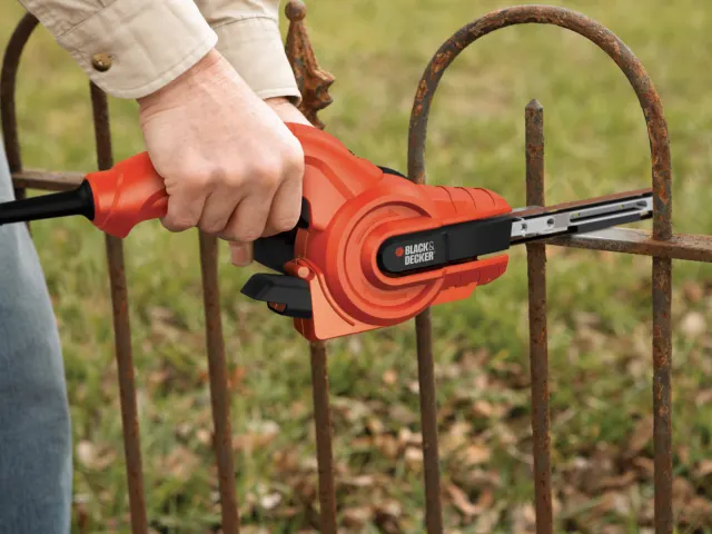 Person using a Black & Decker hedge trimmer on a rusty metal fence with grass in the background