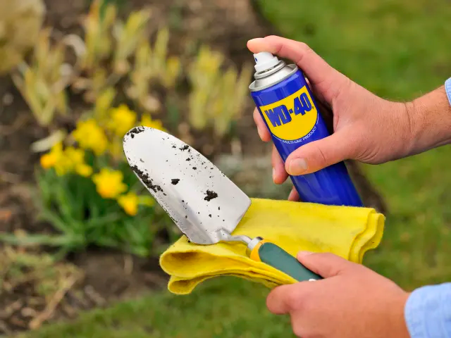 Person cleaning a garden trowel with a yellow cloth and WD-40 spray, outdoors.