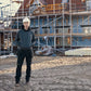 Person wearing a hard hat and dark clothing standing on a construction site with scaffolding and building materials in the background.