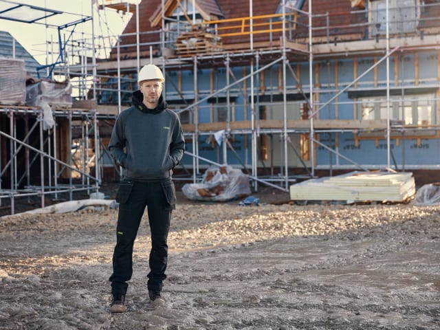 Person wearing a hard hat and dark clothing standing on a construction site with scaffolding and building materials in the background.