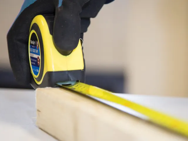 Person measuring a wooden block with a tape measure on a blurred background
