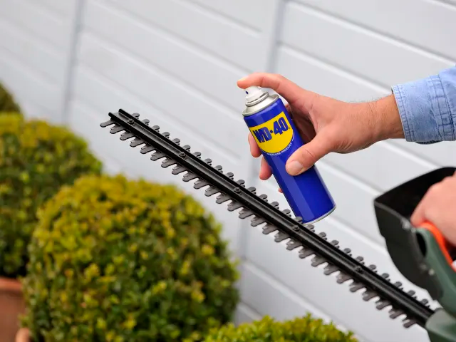 Person using a spray can to clean a hedge trimmer against a white wall.
