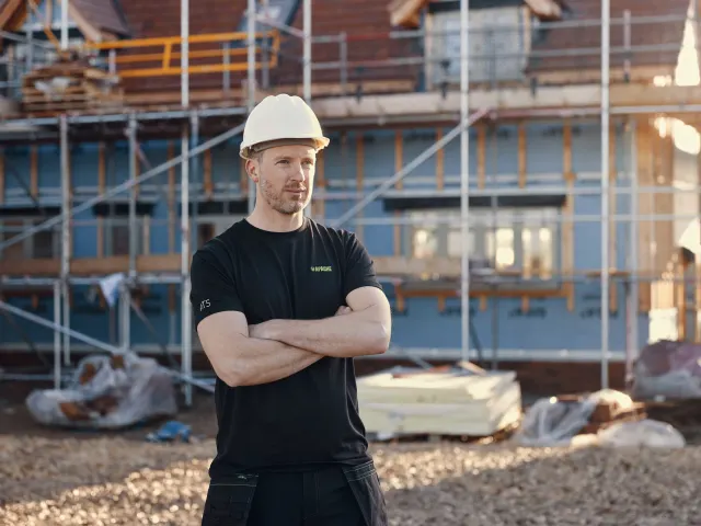 Man wearing a hard hat and black shirt standing in front of a construction site.