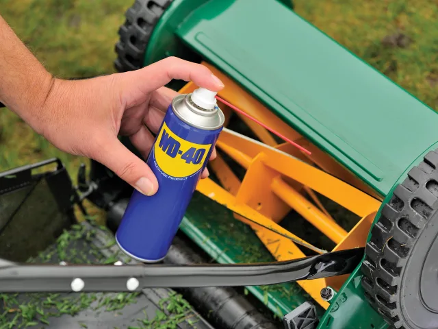 Hand holding a WD-40 spray can near a lawn mower on grass.
