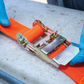 Orange ratchet strap with a metal buckle being adjusted by hands wearing blue gloves on a concrete surface.