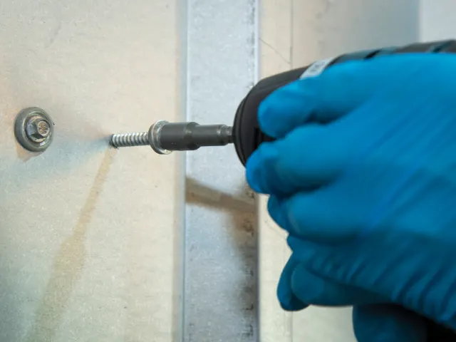 Hand using a drill with a screwdriver bit to tighten a screw into a wall.