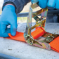Person securing an orange strap with a ratchet to a surface outdoors.