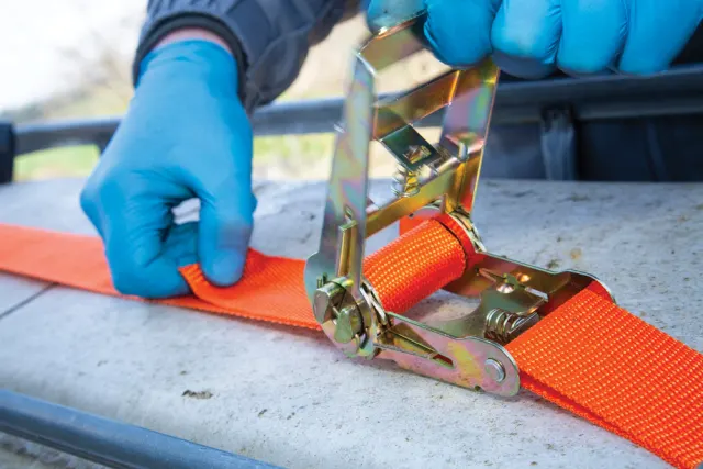Person securing an orange strap with a ratchet to a surface outdoors.