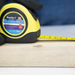 Yellow tape measure with Blue Spot branding on a wooden block, held by gloved hands.
