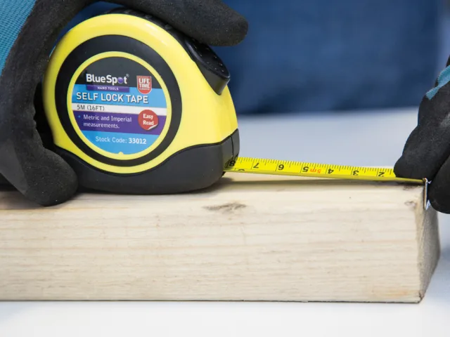 Yellow tape measure with Blue Spot branding on a wooden block, held by gloved hands.